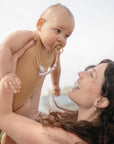 A woman with long brown hair smiles up at a baby in a tan onesie using the FRIGG Daisy Natural Rubber Pacifier (Limited Edition), holding the baby up outdoors on a bright, sunny day.