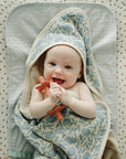Smiling baby on Mushie Changing Pad Liners, wrapped in floral towel and holding a teether.
