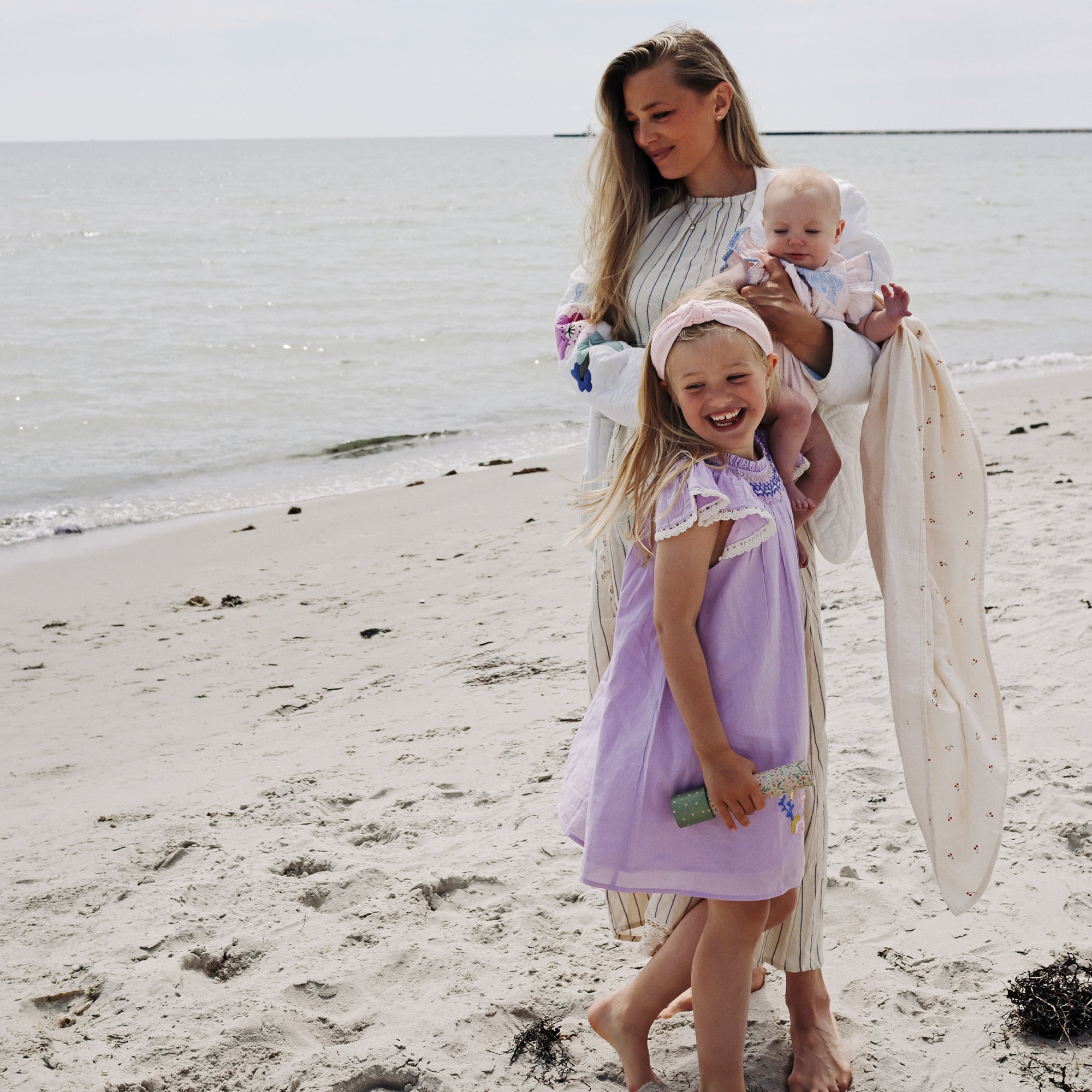 A woman holds a baby in a mushie Organic Cotton Muslin Swaddle Blanket on a sandy beach.