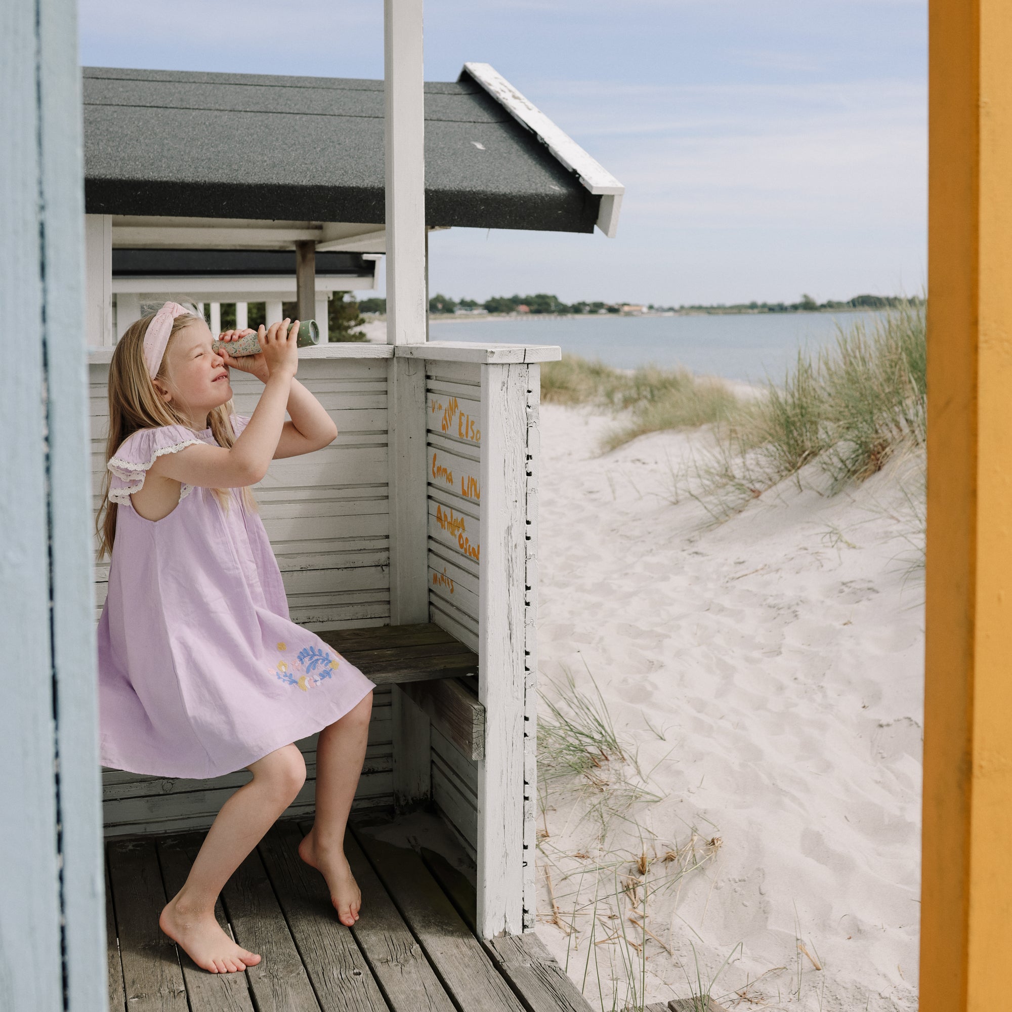 Young girl in lavender dress explores with mushie Kaleidoscopes by sandy dunes.