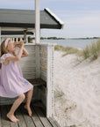 Young girl in lavender dress explores with mushie Kaleidoscopes by sandy dunes.