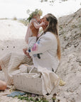 Woman kisses baby on sand, mushie Diaper Caddy by her side.