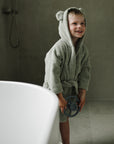 Smiling child in a mushie Bear Robe stands by a bathtub, holding a toy.