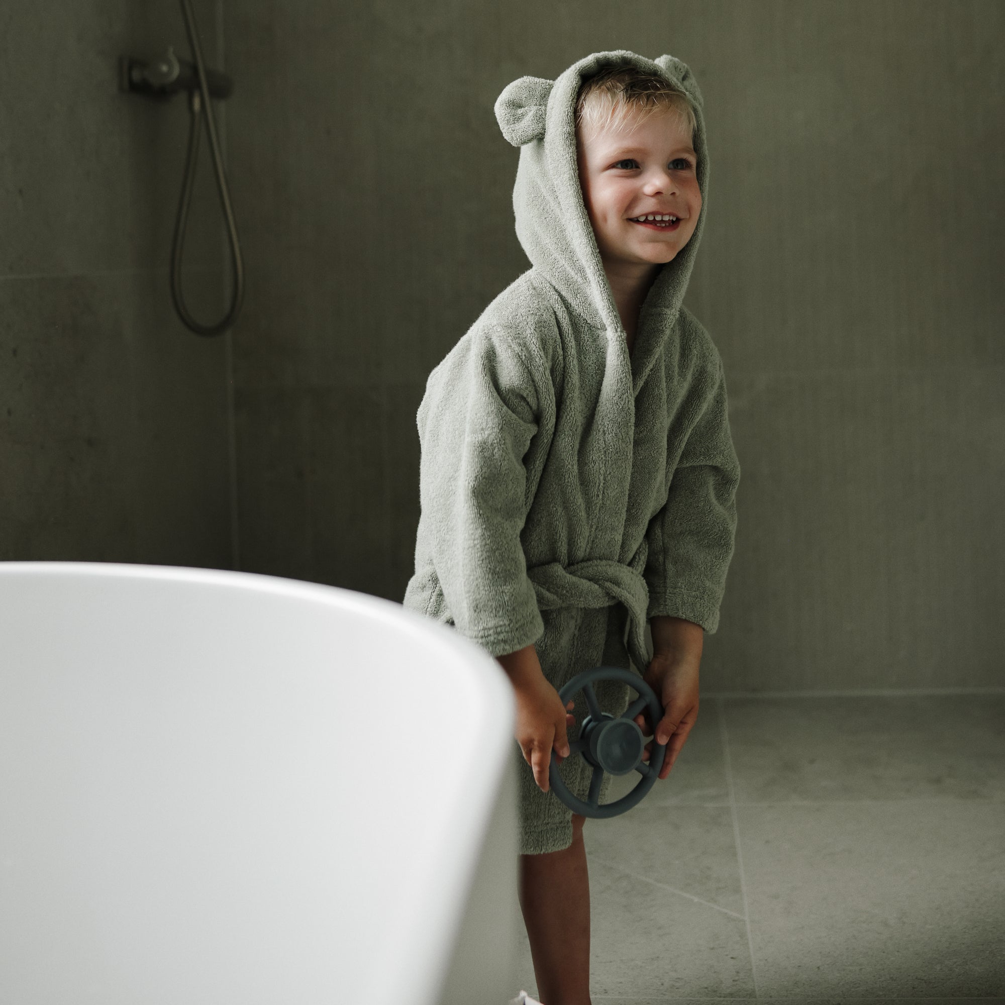 Smiling child holds mushie Suction Spinner Toys by the tub, wearing a bear-eared bathrobe.