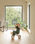A young child rides a mushie Mini Toddler Bike, scanning the room with binoculars.