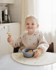 Smiling toddler wearing a mushie Long Sleeve Bib while eating at a table.