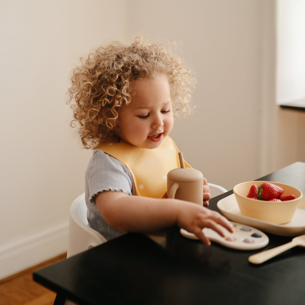 A toddler wearing a mushie Silicone Baby Bib sits at a table with a bowl of strawberries.