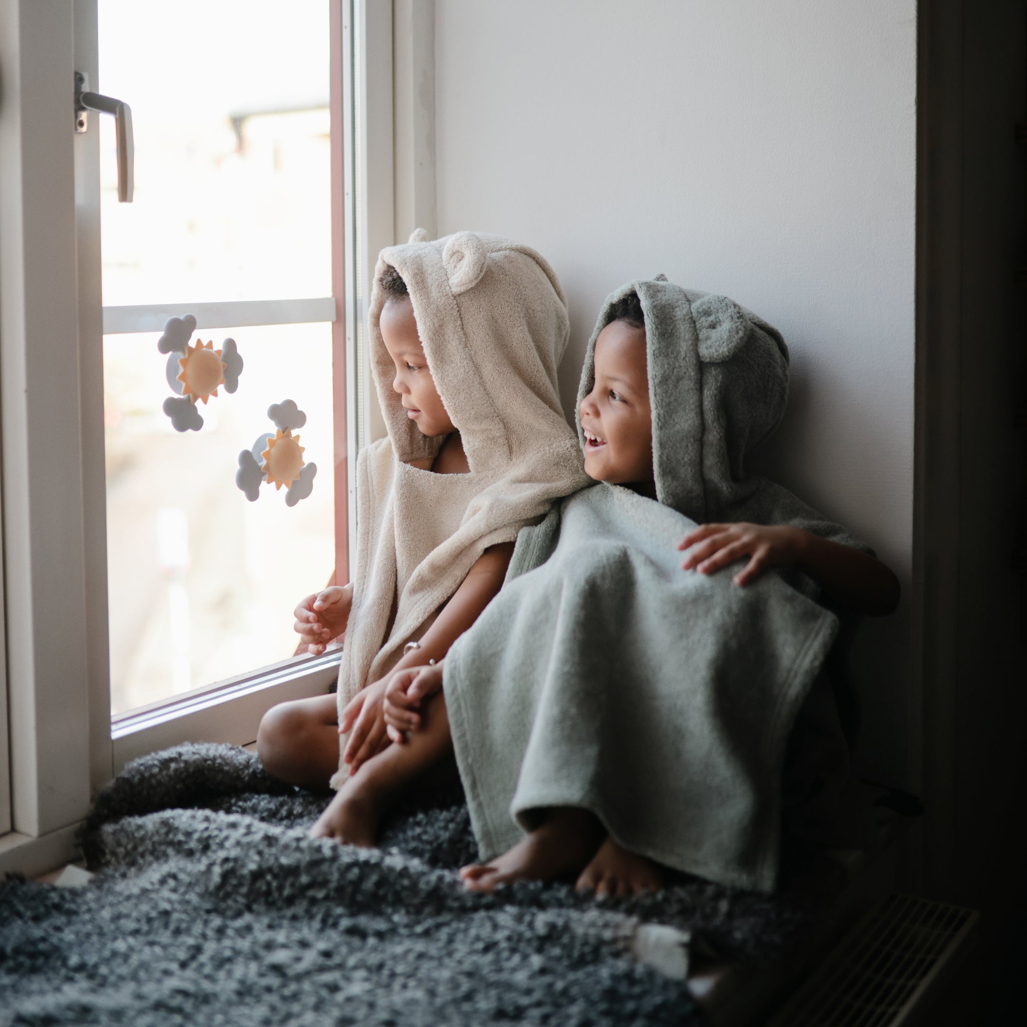 Two kids in mushie Bear Poncho Towels smile by a window, wearing cozy organic cotton.