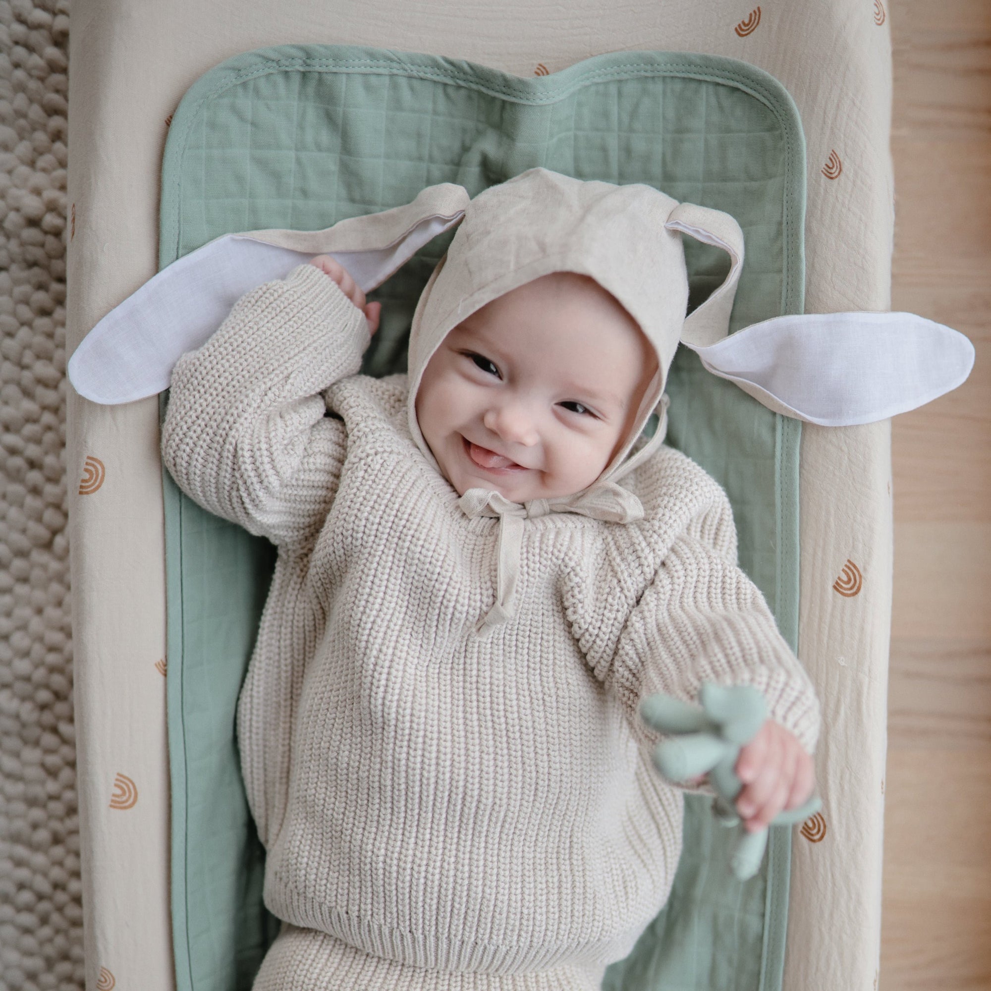 Happy baby in Mushie Changing Pad Liners and organic cotton knit suit with bunny-ear hat.
