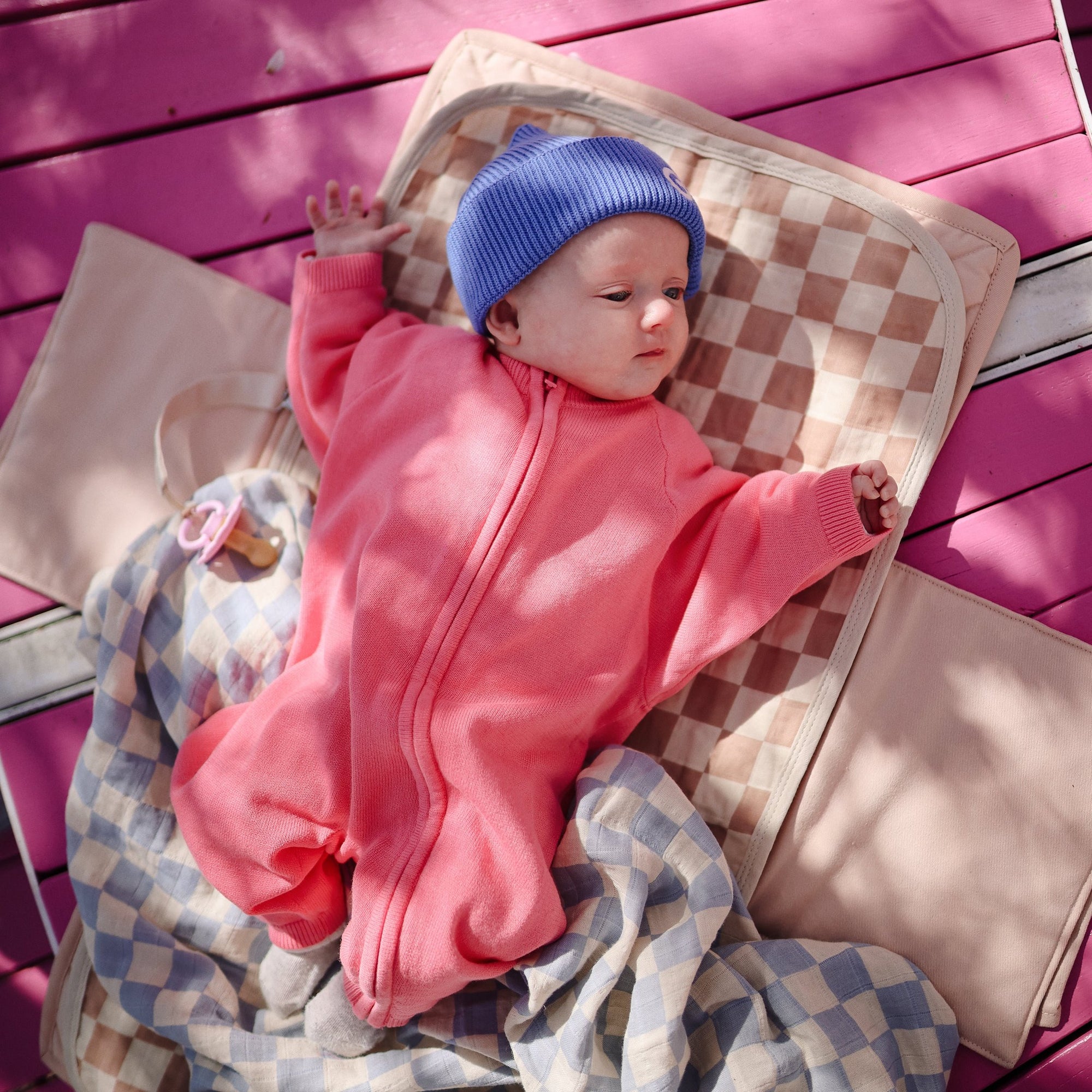 Baby in pink suit on Mushie Changing Pad Liner, blue hat, pink background.