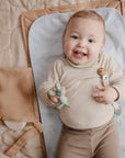 Smiling baby on Mushie Changing Pad Liner holds a toy dinosaur while lying on the mat.