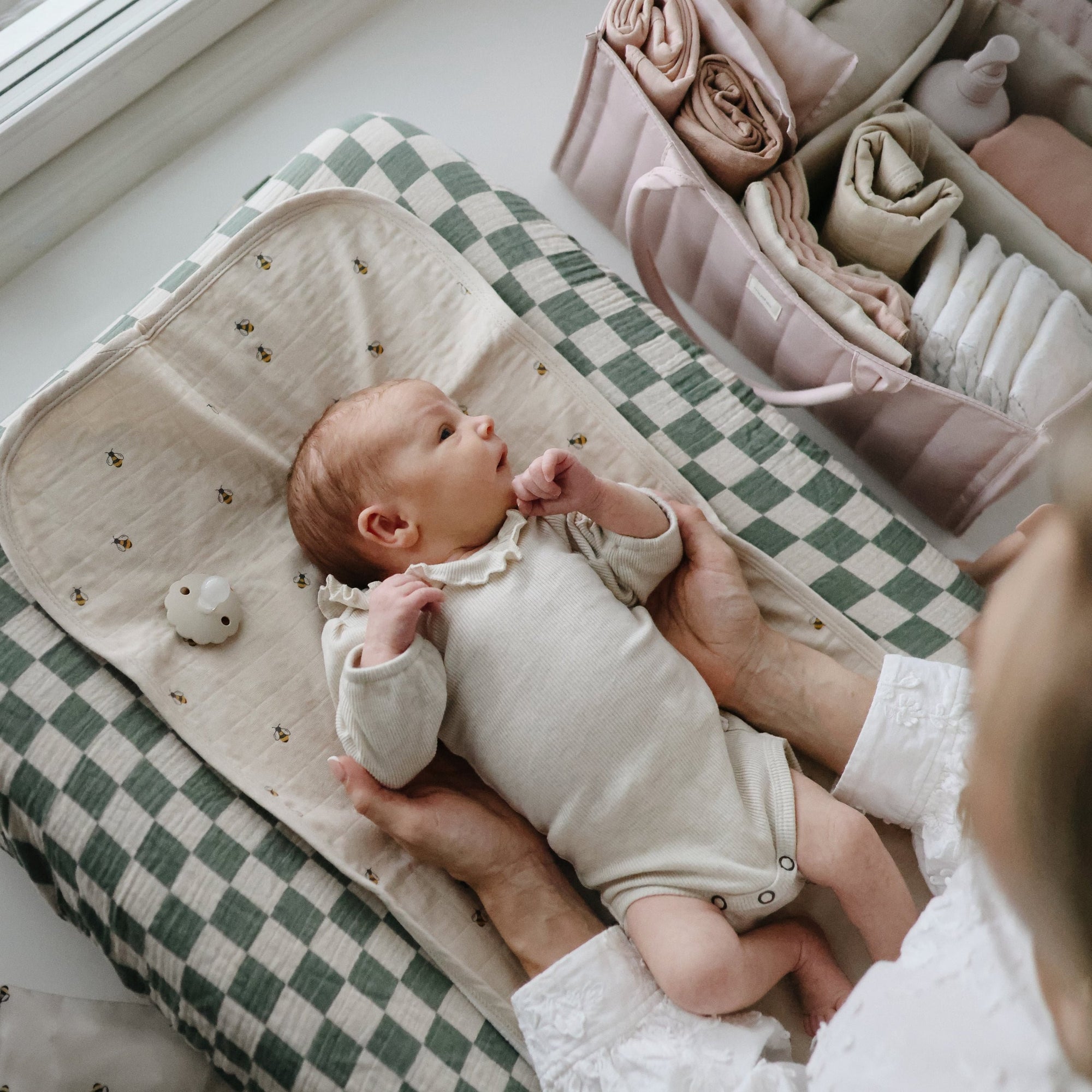 Baby in cream onesie on Mushie Changing Pad Liner, held gently near a stack of folded linens.
