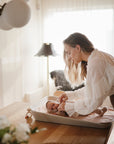 A woman uses Mushie Changing Pad Liners to change a baby’s diaper on a table.