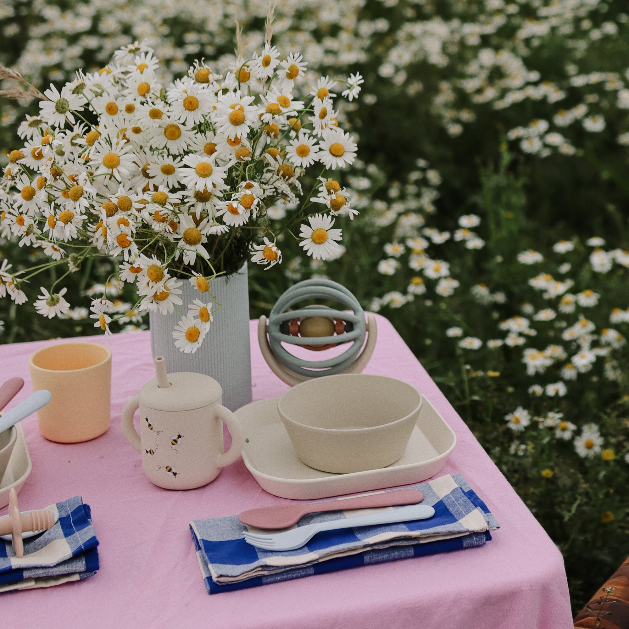 A table with daisies, dishes, mushie Dinnerware Cups, and napkins on a pink cloth outdoors.