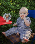 Smiling baby in checkered outfit with FRIGG Daisy Silicone Pacifier by a red table outdoors.