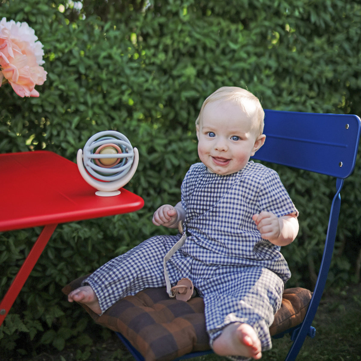 Baby with mushie Linen Pacifier Clip 2-Pack sits by red table with toy, outdoors.