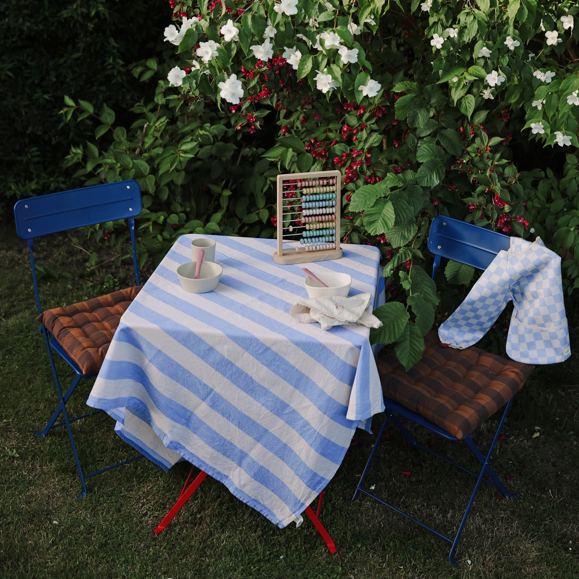 Long sleeve bib by mushie on a table with abacus and bowls near flowering bushes.
