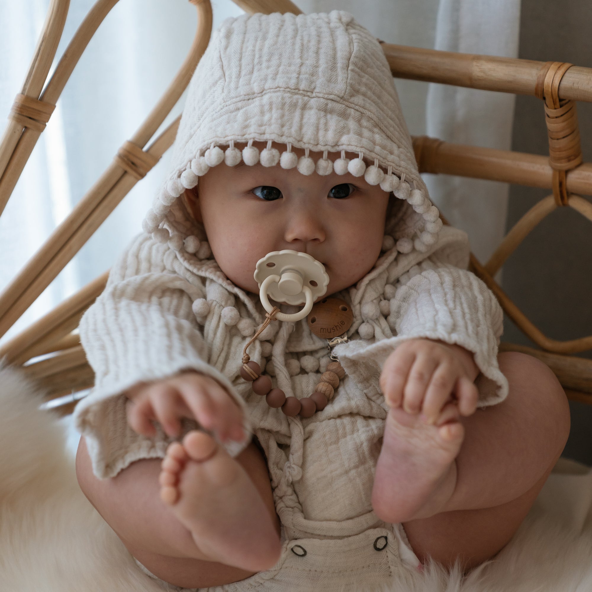 A baby sits on a chair in a textured cream hooded outfit with pom-pom trim, holding their feet and using the FRIGG Daisy Silicone Pacifier 2-Pack | Limited Edition for comfort, attached to a beaded clip.