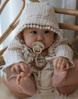 A baby sits on a chair in a textured cream hooded outfit with pom-pom trim, holding their feet and using the FRIGG Daisy Silicone Pacifier 2-Pack | Limited Edition for comfort, attached to a beaded clip.