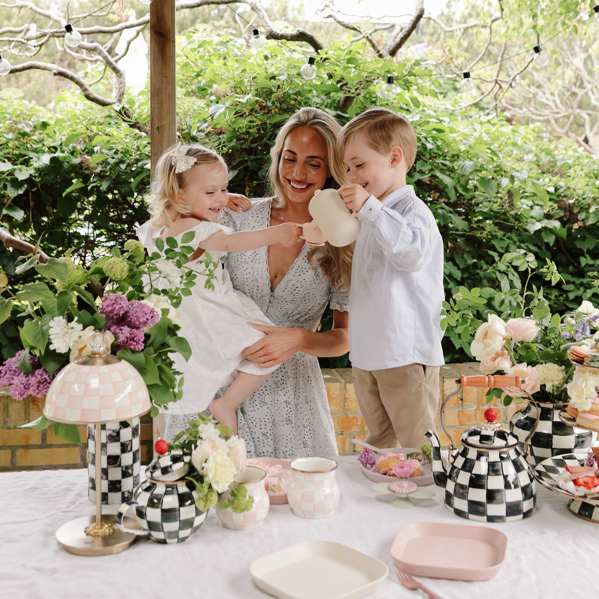 A woman and two kids pour tea from a Mushie Silicone Tea Play Set at a flower-decorated table.