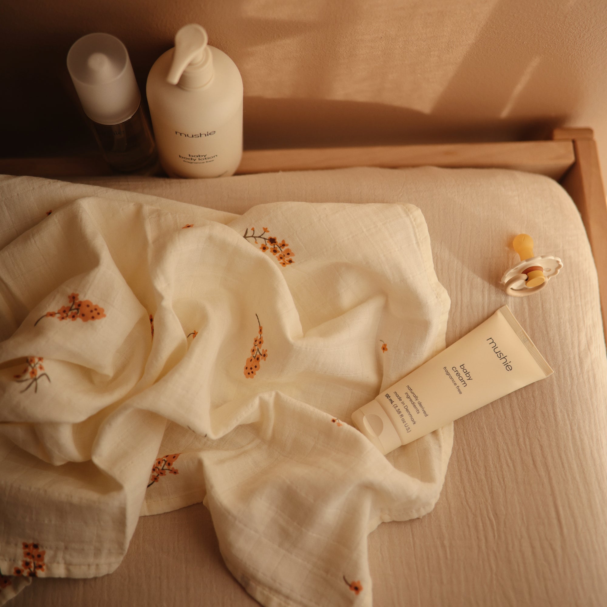 Mushie Baby Cream and a pacifier on a beige surface with a cream floral baby blanket.