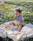 A toddler sits on a mushie Organic Cotton Muslin Swaddle Blanket, snacking outdoors in nature.
