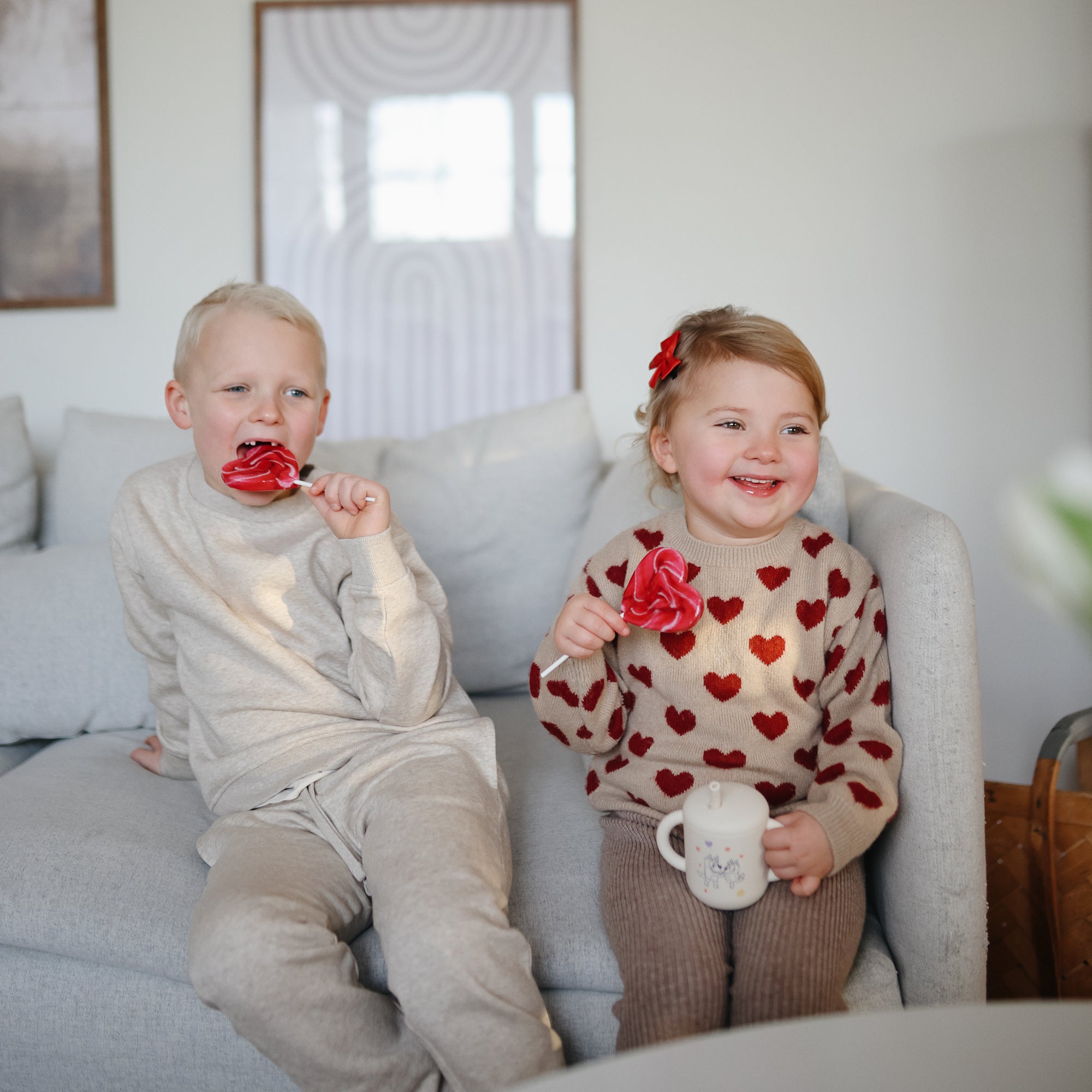A young boy and girl sit on a light couch. The boy wears beige and holds a red heart lollipop, while the smiling girl in a red-heart sweater holds a mug. Nearby is her Mushie Bluey x Mushie Silicone Training Cup + Straw, made of food-grade silicone.