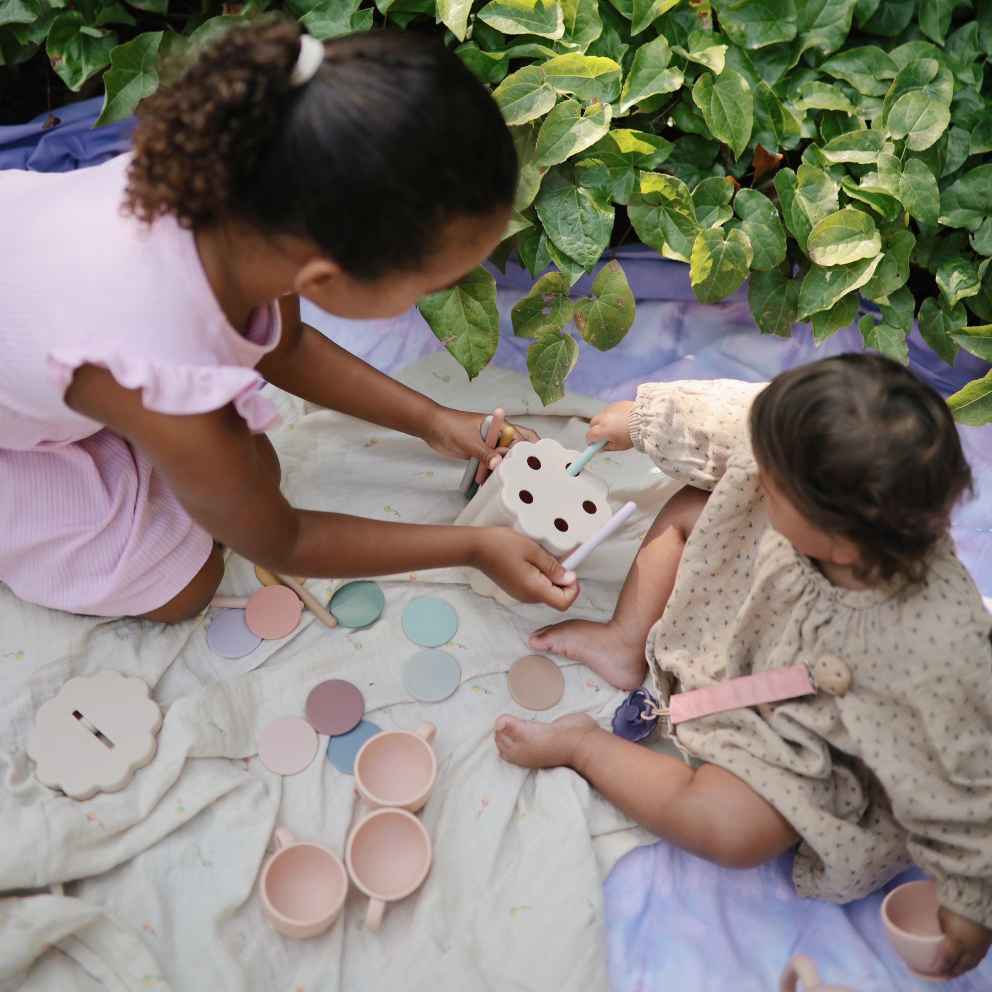 Two children play on a mushie Organic Cotton Muslin Swaddle Blanket near green foliage.