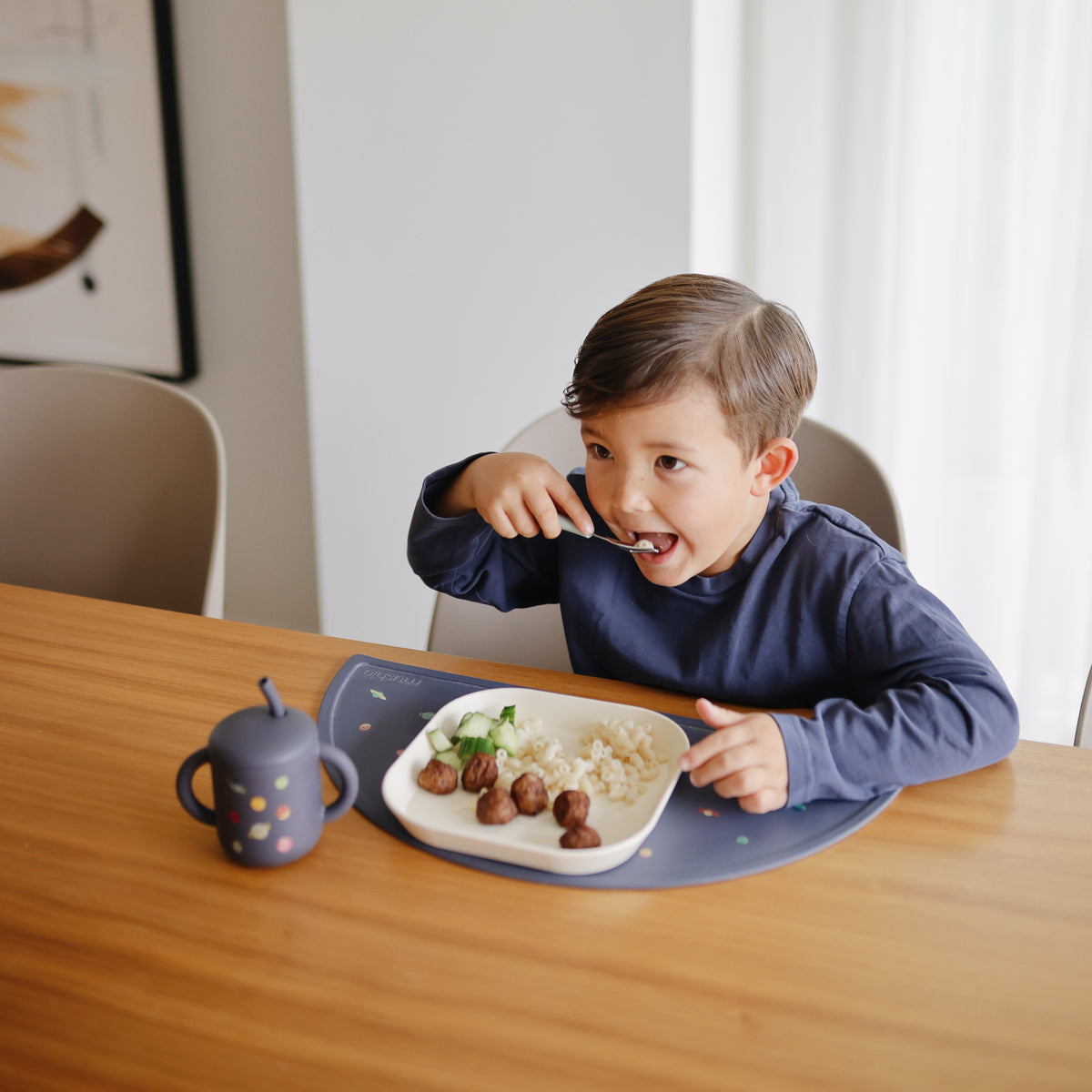 A child eats at a table next to a Mushie Silicone Training Cup + Straw.