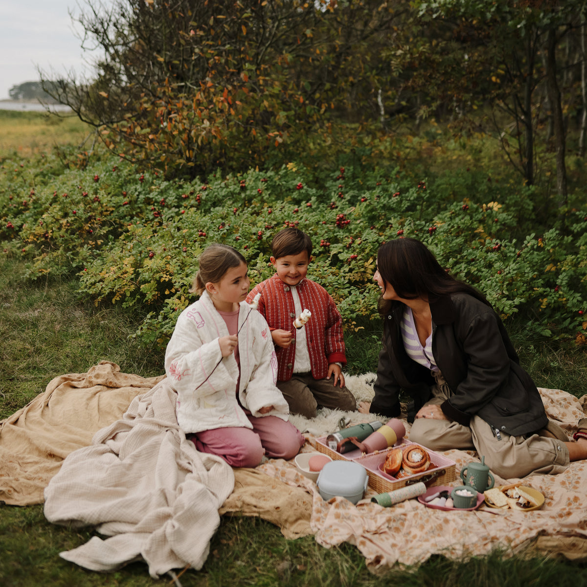 A woman and two children sit outdoors with mushie Kaleidoscopes, eating marshmallows on sticks.