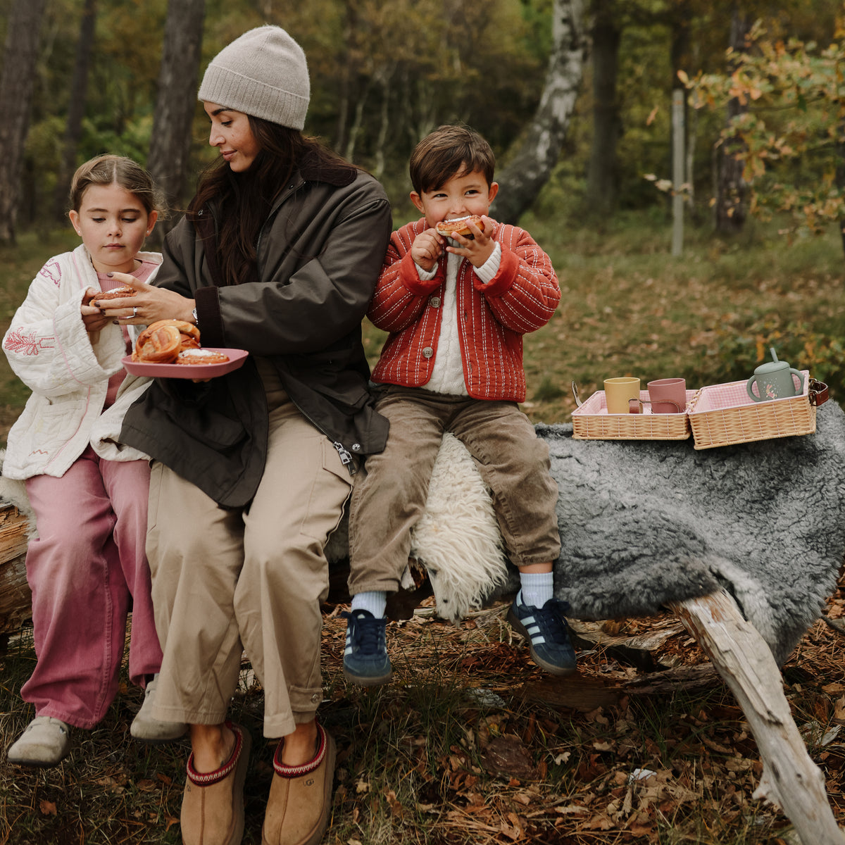 A woman and two children sit on a fallen tree, eating pastries with a Mushie Silicone Training Cup.