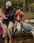 A woman and two children sit on a fallen tree, eating pastries with a Mushie Silicone Training Cup.