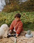 A child sits on an outdoor blanket with a small white dog and a mushie lunchbox.