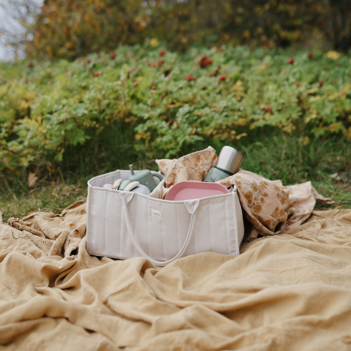 A quilted mushie Diaper Caddy filled with baby items, sitting on a blanket outdoors.