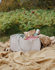 A quilted mushie Diaper Caddy filled with baby items, sitting on a blanket outdoors.