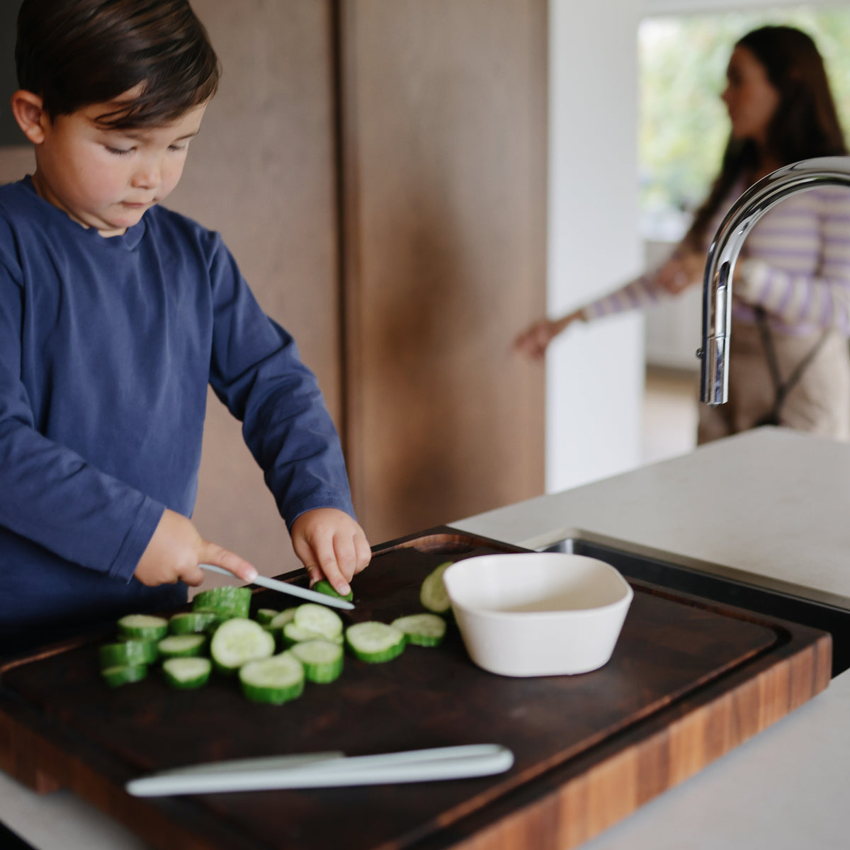 A child slices cucumbers on a wooden board next to an empty mushie Square Dinnerware Bowl.