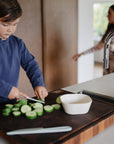 A child slices cucumbers on a wooden board next to an empty mushie Square Dinnerware Bowl.