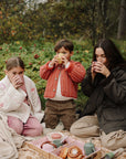 A mother and two children sit on a picnic blanket drinking from Mushie silicone food containers.