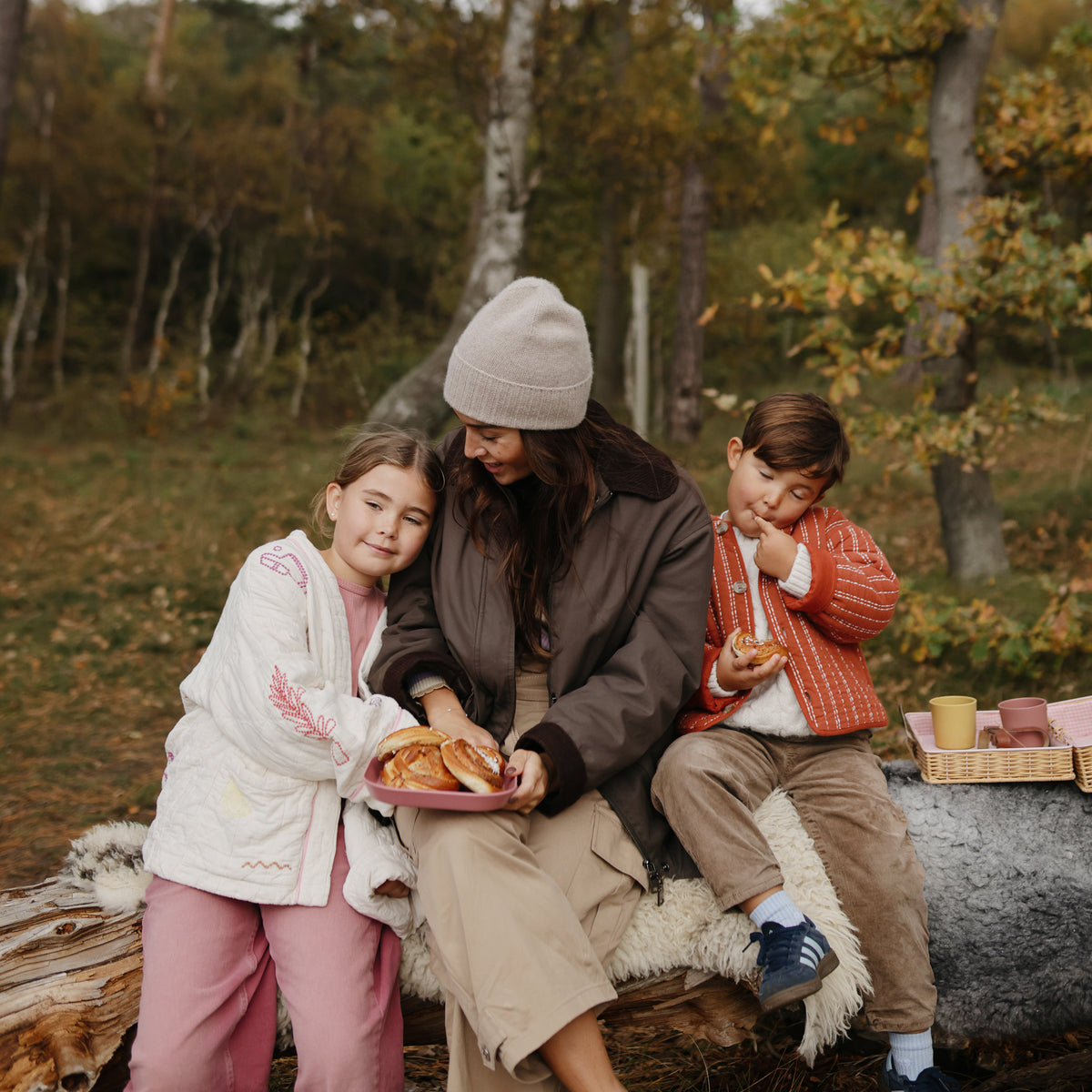 A woman and two children sit on a log, sharing pastries from a mushie Square Dinnerware Plate.