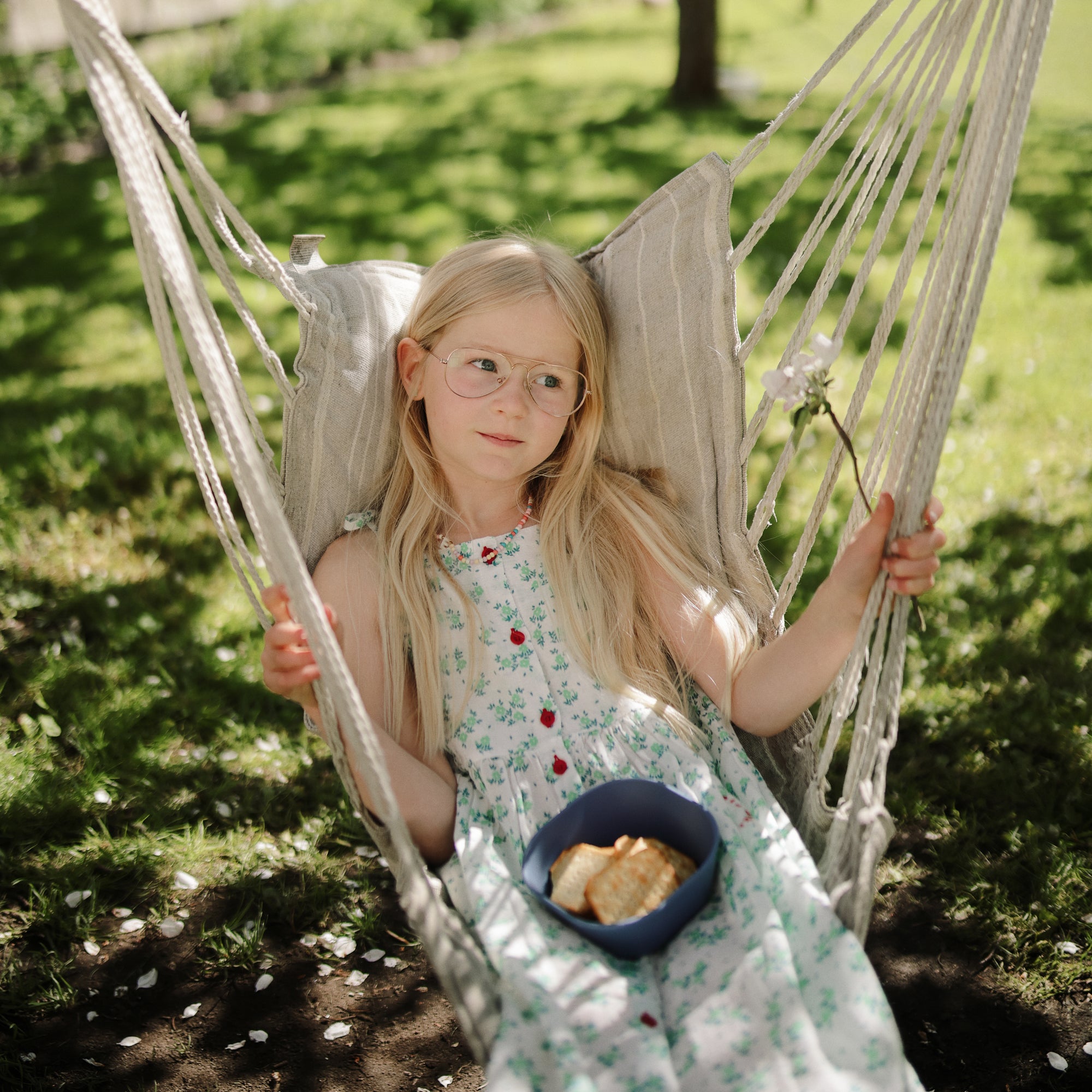 Girl with long blonde hair holds mushie Square Dinnerware Bowl in hammock, wearing glasses.