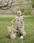 A toddler in a strawberry dress holds a mushie Linen Pacifier Clip 2-Pack by a beige tricycle.