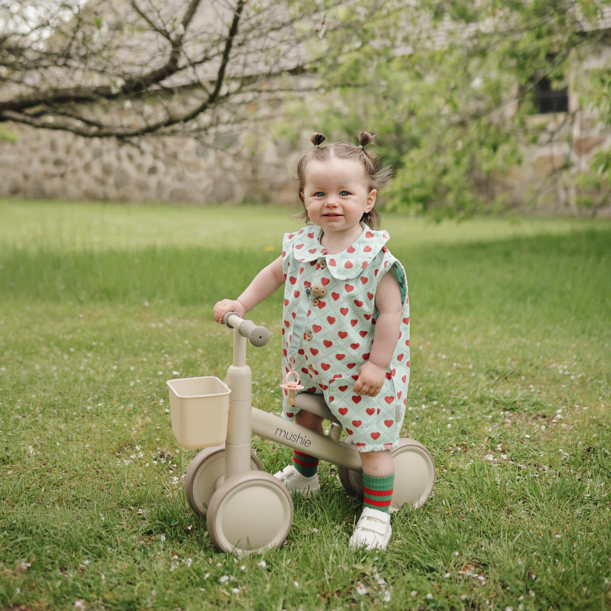 Toddler in a strawberry dress stands with a mushie Mini Toddler Bike on grass outdoors.