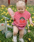 Toddler with mushie Linen Pacifier Clip 2-Pack sits on a balance bike among dandelions.
