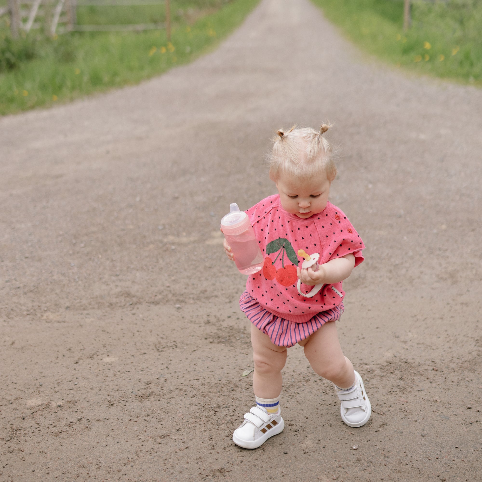 Toddler with mushie Linen Pacifier Clip 2-Pack in pink outfit holding a snack on dirt road.