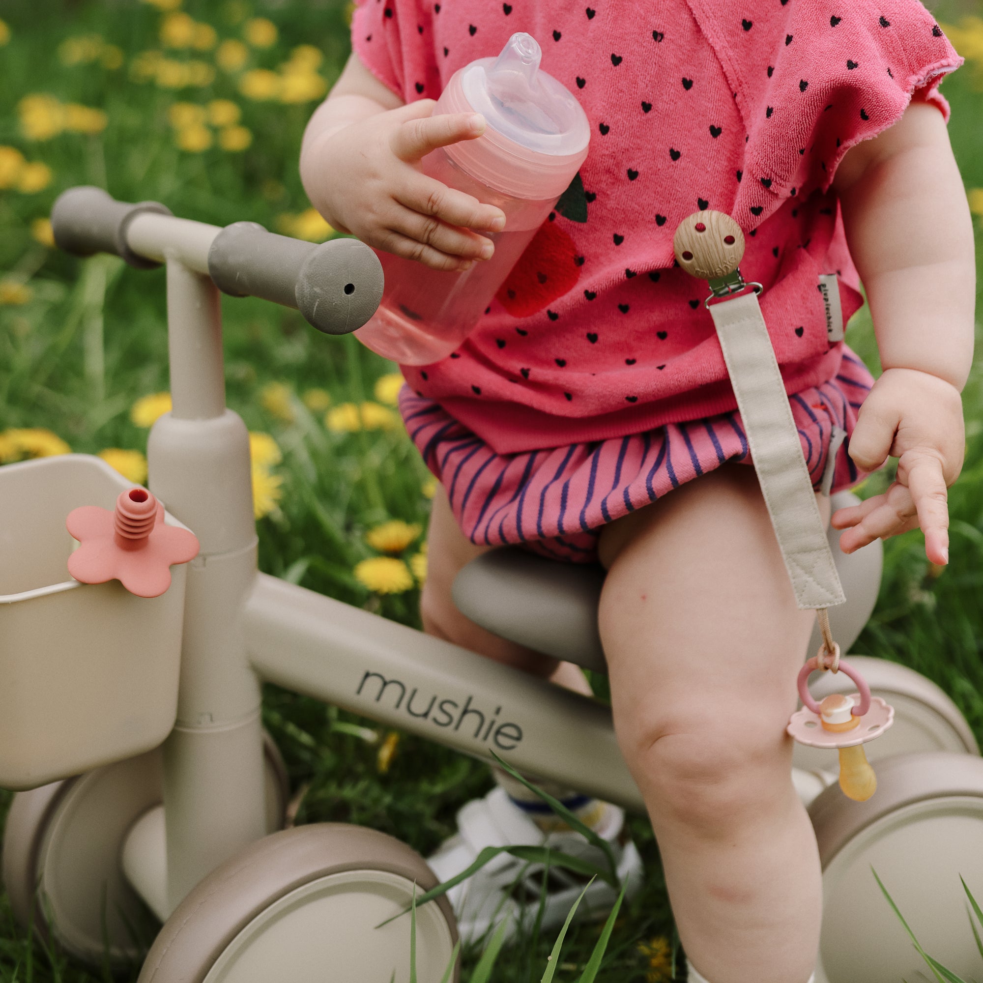A toddler with a mushie Linen Pacifier Clip 2-Pack on beige tricycle, sitting on green grass.