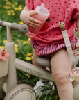 A toddler with a mushie Linen Pacifier Clip 2-Pack on beige tricycle, sitting on green grass.