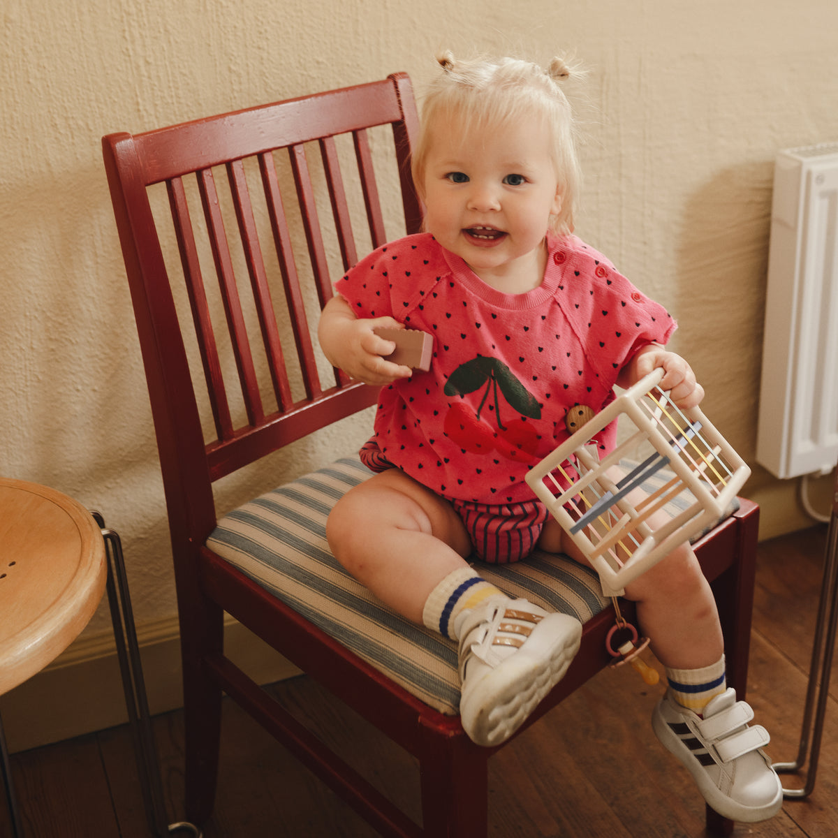 A toddler sits on a chair holding a mushie Elastic Shape Sorter to practice fine motor skills.