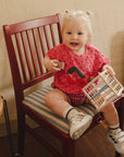 A toddler sits on a chair holding a mushie Elastic Shape Sorter to practice fine motor skills.
