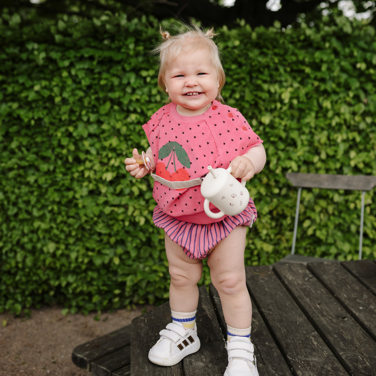 Happy toddler in pink with mushie Linen Pacifier Clip 2-Pack stands holding a sippy cup outside.