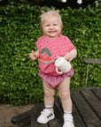 Happy toddler in pink with mushie Linen Pacifier Clip 2-Pack stands holding a sippy cup outside.